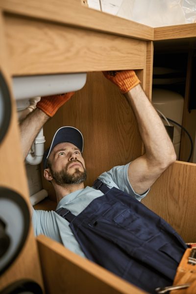 Bearded young man handyman wearing work gloves and cap while repairing sink
